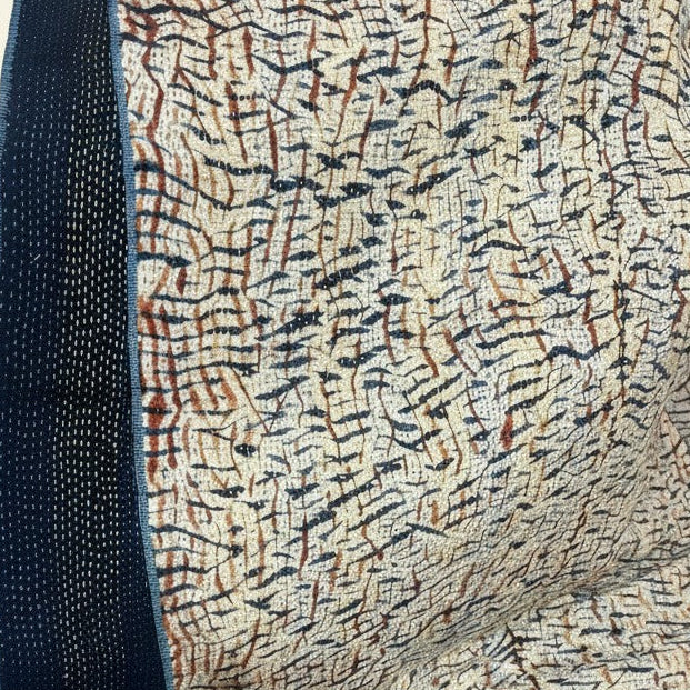 A close-up of a patterned textile with a cream base, dense black and brown linear motifs, and a dark navy woven border along the left edge, shown against a light beige background.