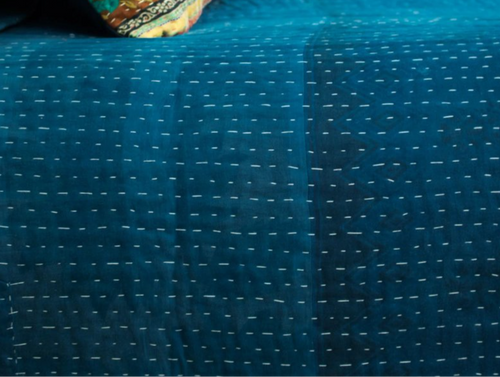A close-up of a teal blue quilted bedspread or duvet with visible white stitching, with a patterned decorative pillow resting on it in a bedroom setting.